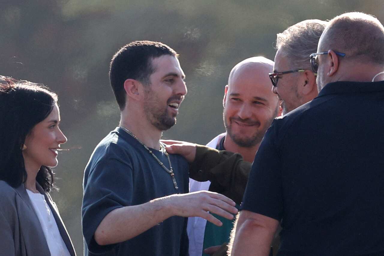 A man wearing a grey shirt smiling, standing near three other men and a woman who are all also smiling.