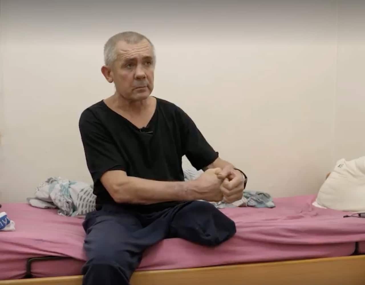 A man in a black top and dark pants sits on a bed. He has one leg cut off above the knee and has a serious expression on his face.