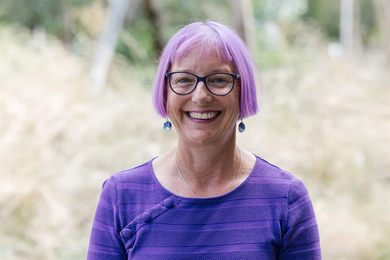 A woman in a purple top and matching hair smiles at the camera while standing outside.