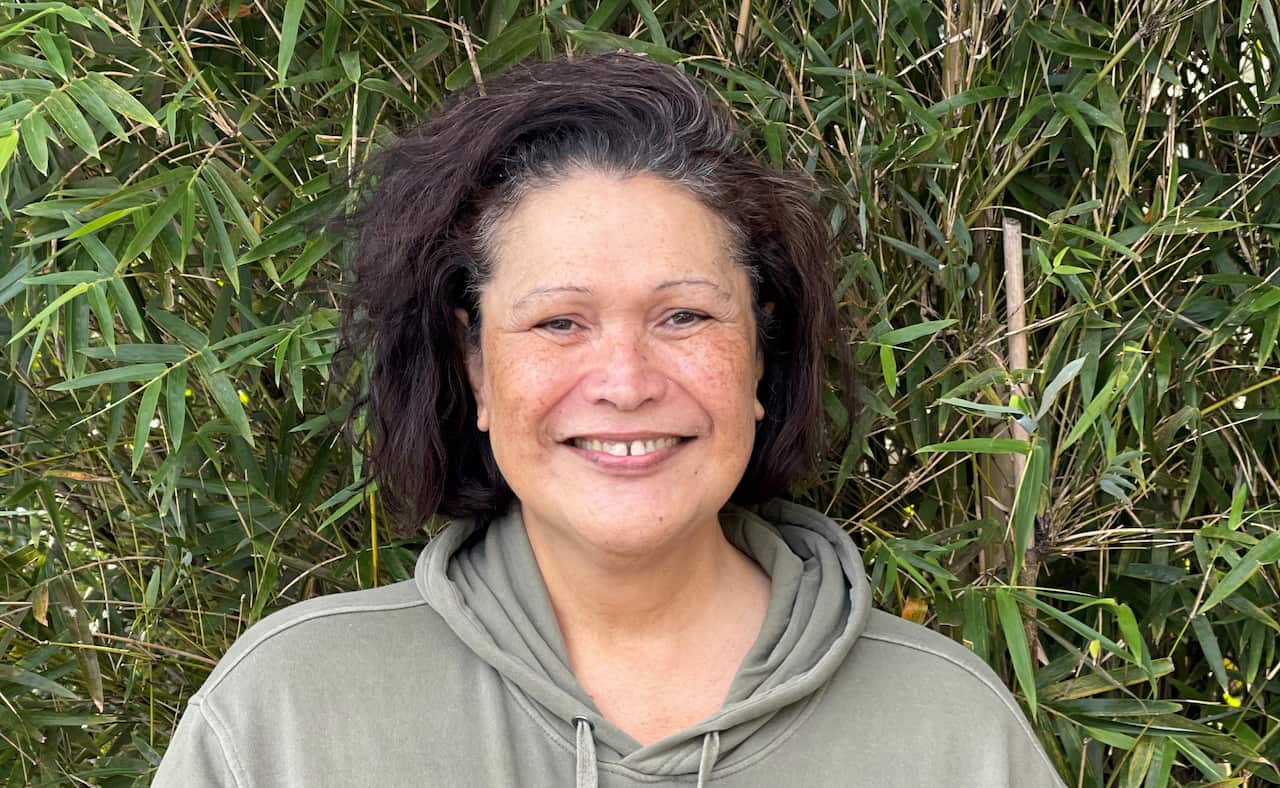 Woman smiles in a green hood jumper in front of greenery.  