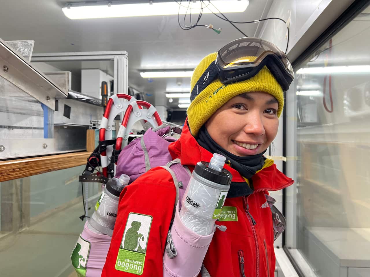 A woman wearing winter clothing, including a beanie and snow goggles on her head, with snowshoes strapped to the backpack on her back.