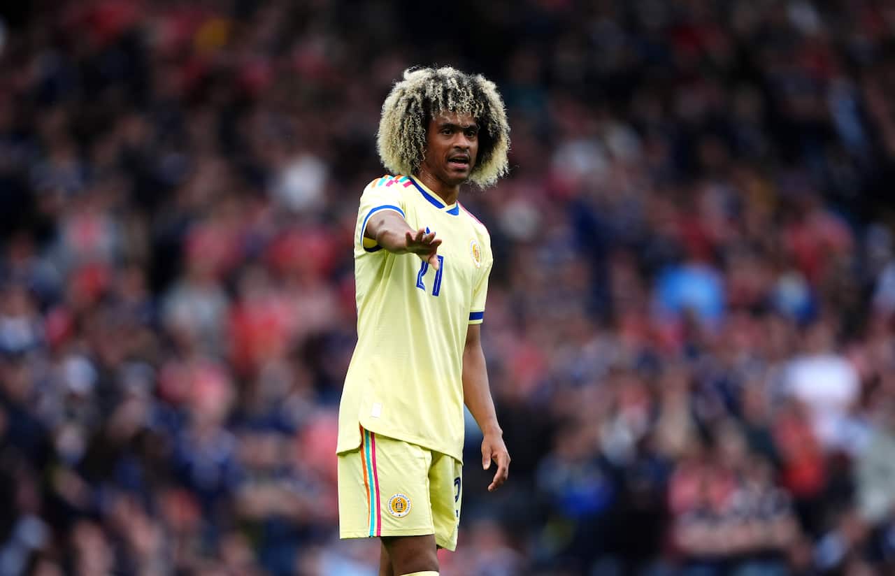 A man in a yellow football kit stands in front of a large crowd, mid game. His arm is outstretched. 