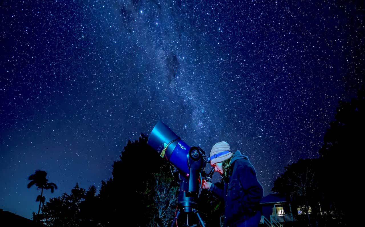 A person looks through a telescope on a dark night. The Milky Way shines brightly, visible to the human eye.