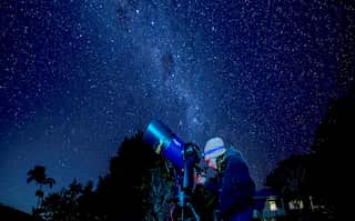 A person looks through a telescope on a dark night. The Milky Way shines brightly, visible to the human eye.