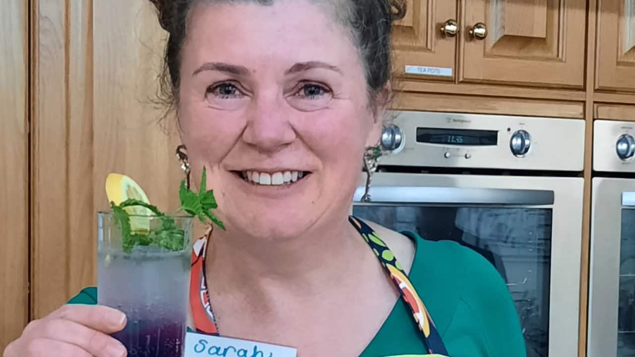 A middle-aged woman in a colourful apron and green shirt smiles to the camera, standing in the kitchen and holding a purple mocktail.