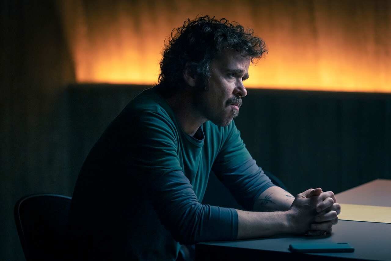 A man with curly hair and a beard, in a long-sleeved t-shirt, leans on a table in a dark room. Behind him, an orange light fills the top half of the far wall. 