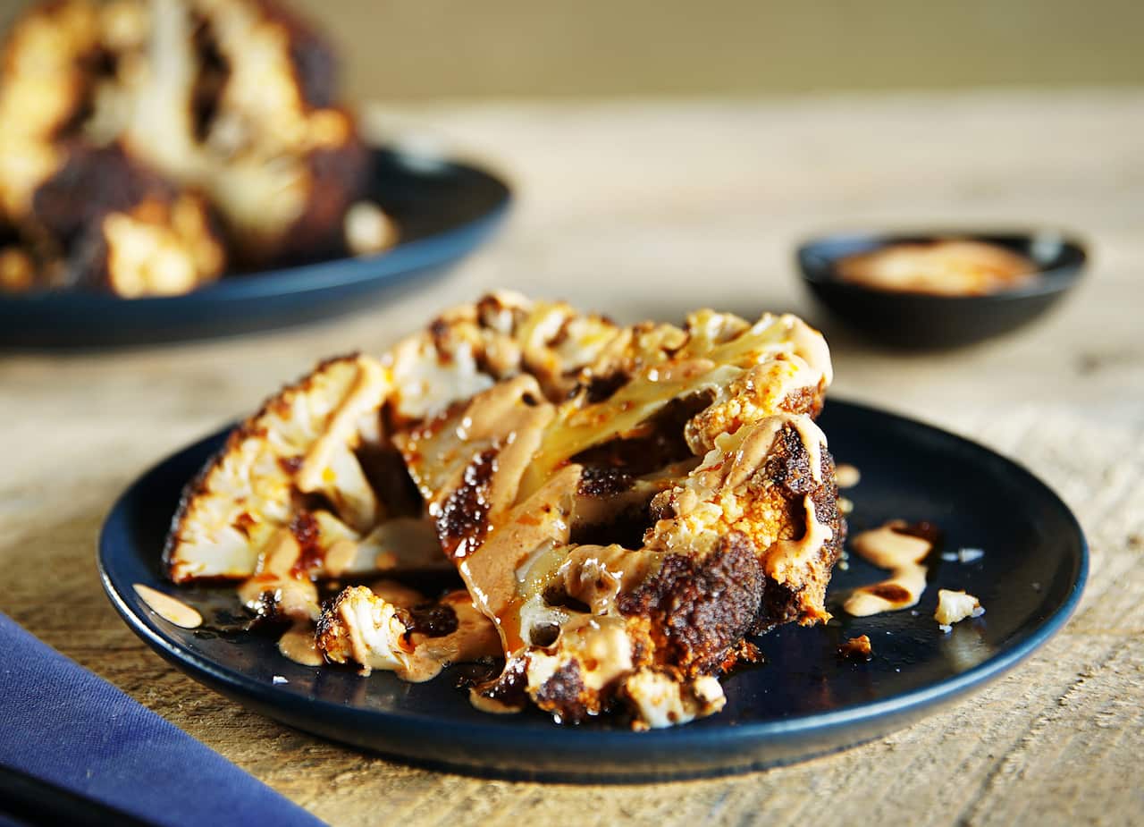 Cooked wedges of cauliflower, with brown edges, and drizzled with sauce, sit on a round dark blue plate.