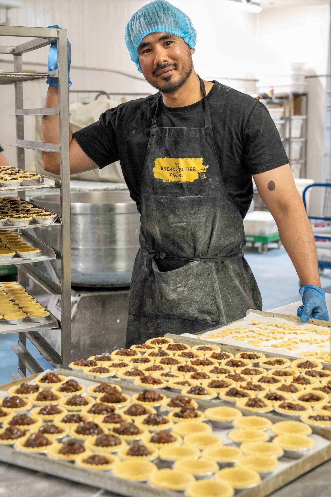 A man in a black t-shirt stands behind a tray of fruit mince pies.