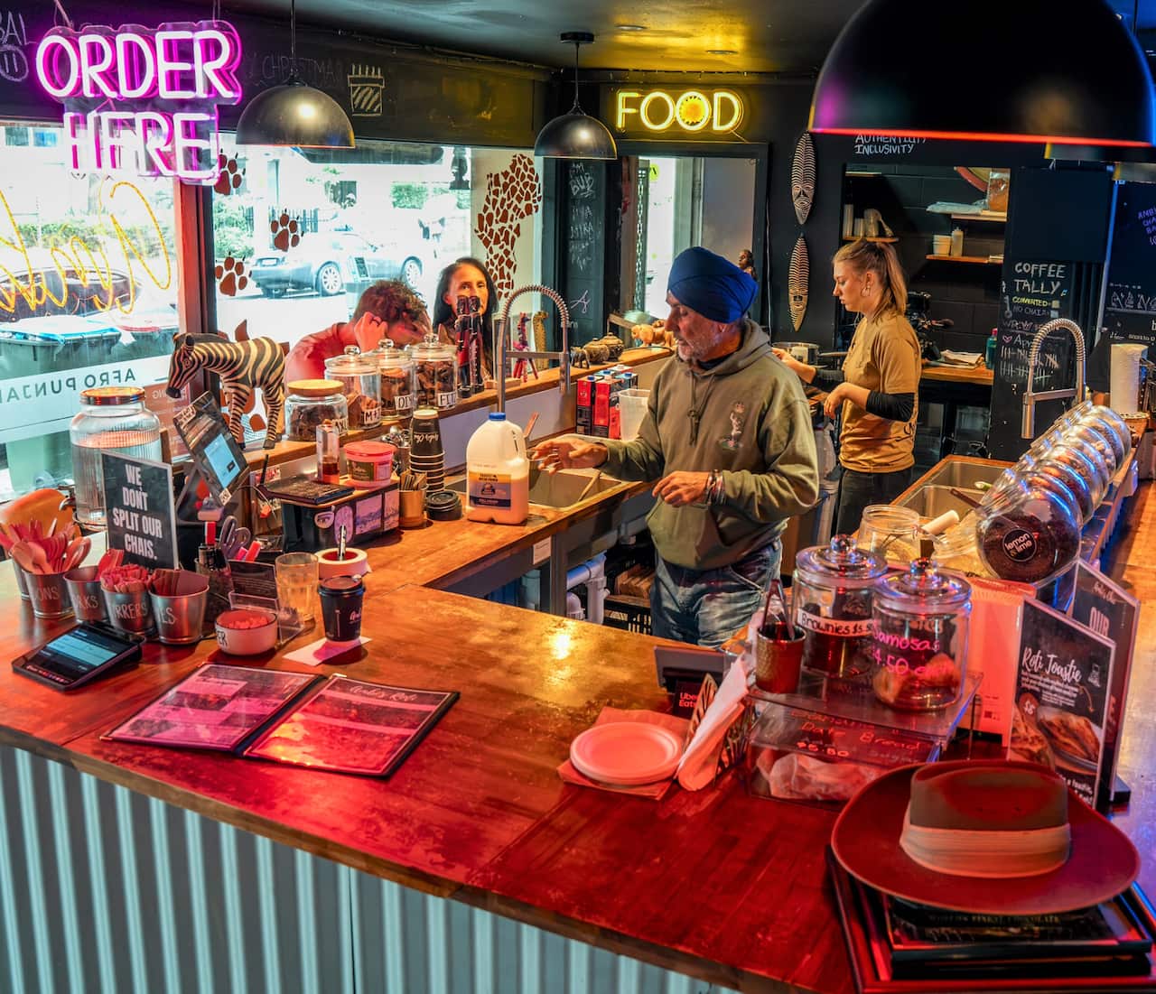 A colourful chai bar with customers sitting at wooden counters while staff make and serve tea.