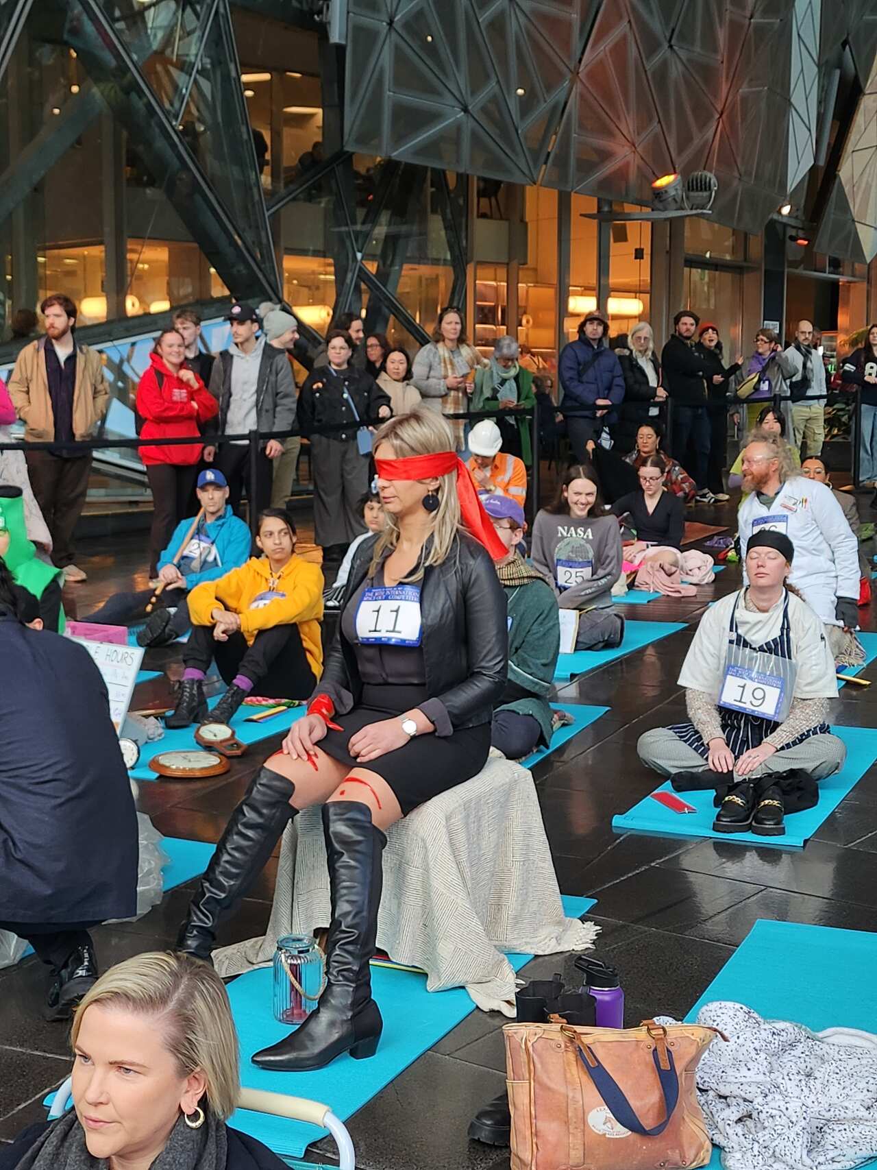 A woman sitting on a stool with a red blindfold surrounded by others sitting on blue mats.