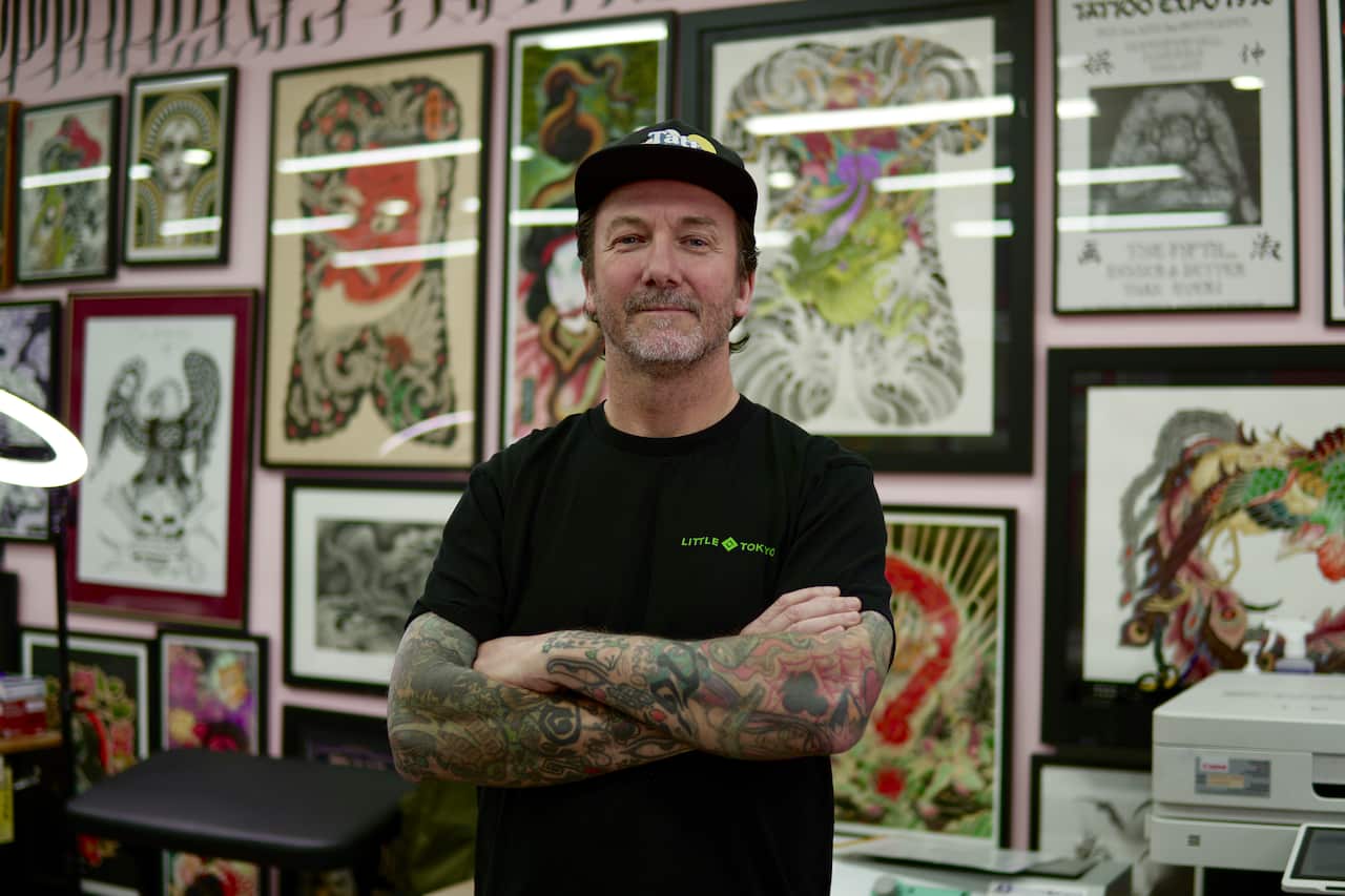 A man in a black T-shirt and cap stands with his tattooed arms crossed inside a tattoo parlour, with framed tattoo pictures in the background.