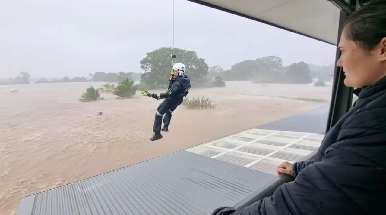 A man abseiling down from a helicopter onto a house roof surrounded by brown floodwater.