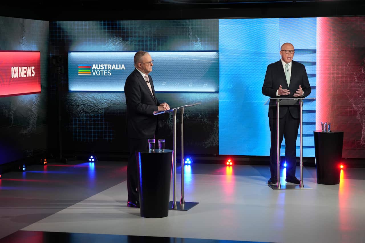 Two men in black suits stand at lecterns placed at a distance in a news studio. There are screens lit in red and blue colours behind them.