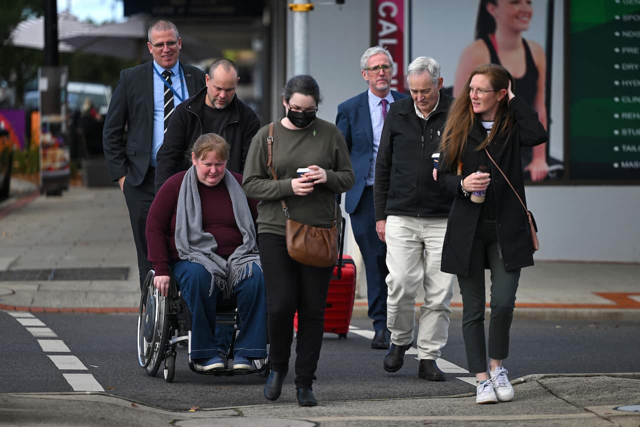 A small group of people crossing a road. One woman is in a wheelchair.