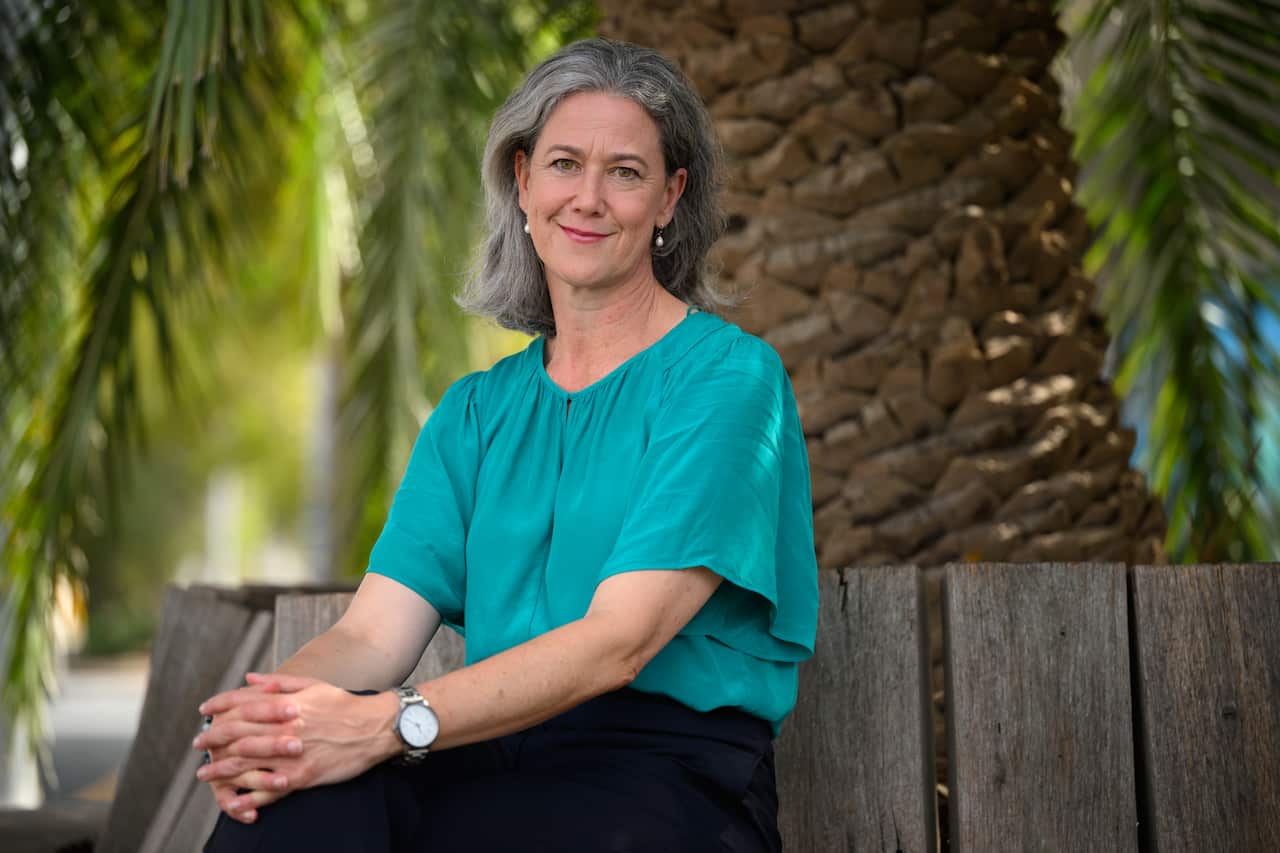 A woman in a turquoise shirt with silver hair smiles as she sits on a bench outside.