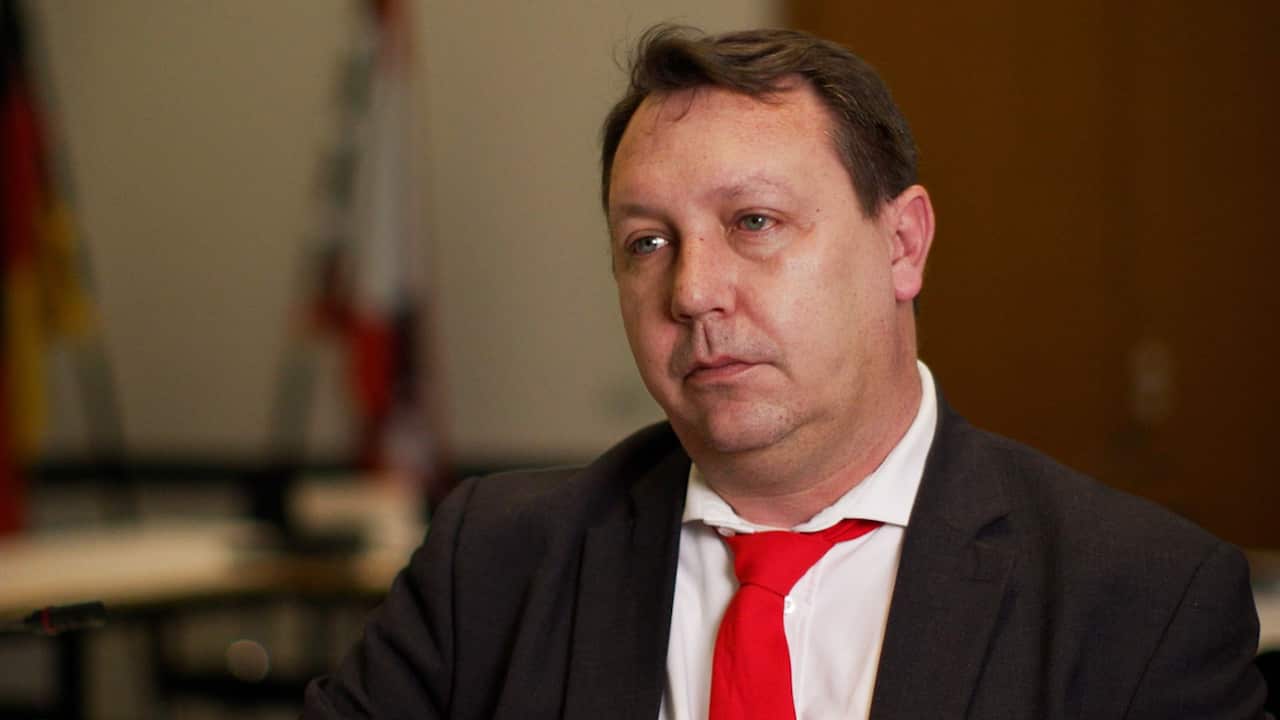 A middle-aged man in a back suit, white shirt and red tie.