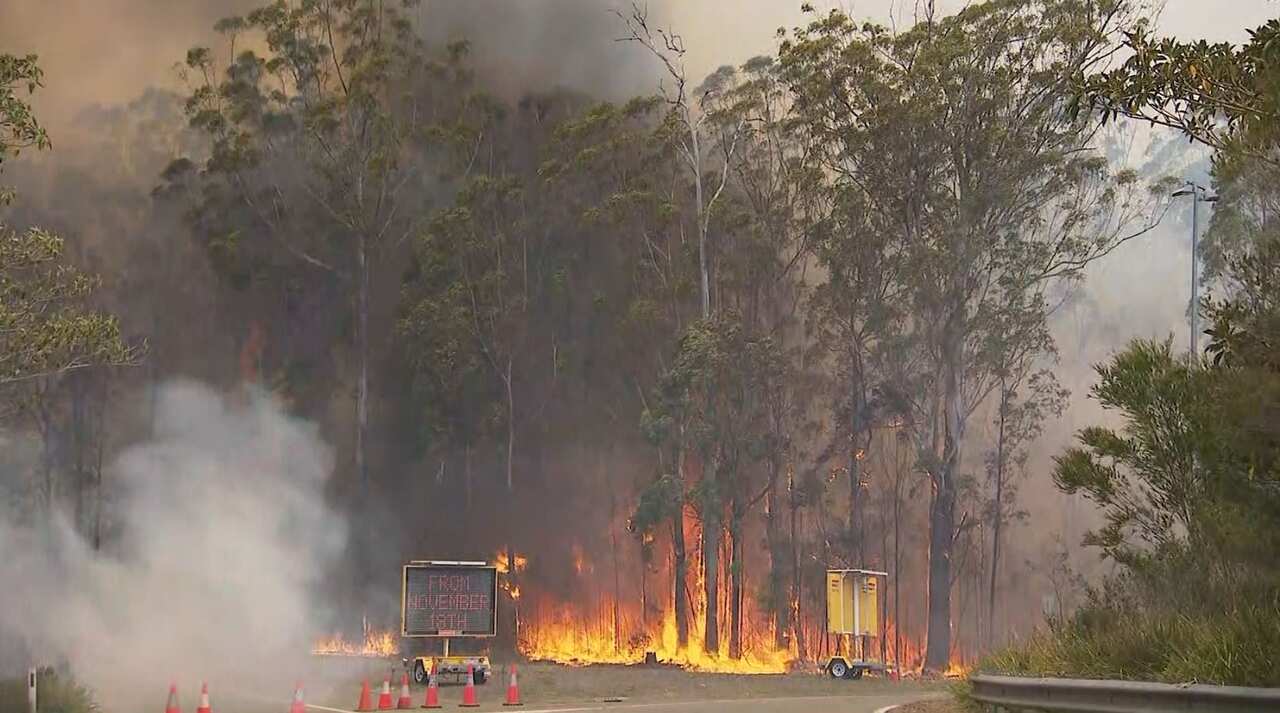 A bushfire rages as it reaches the road, where an electronic sign reads 'From November 18th'.