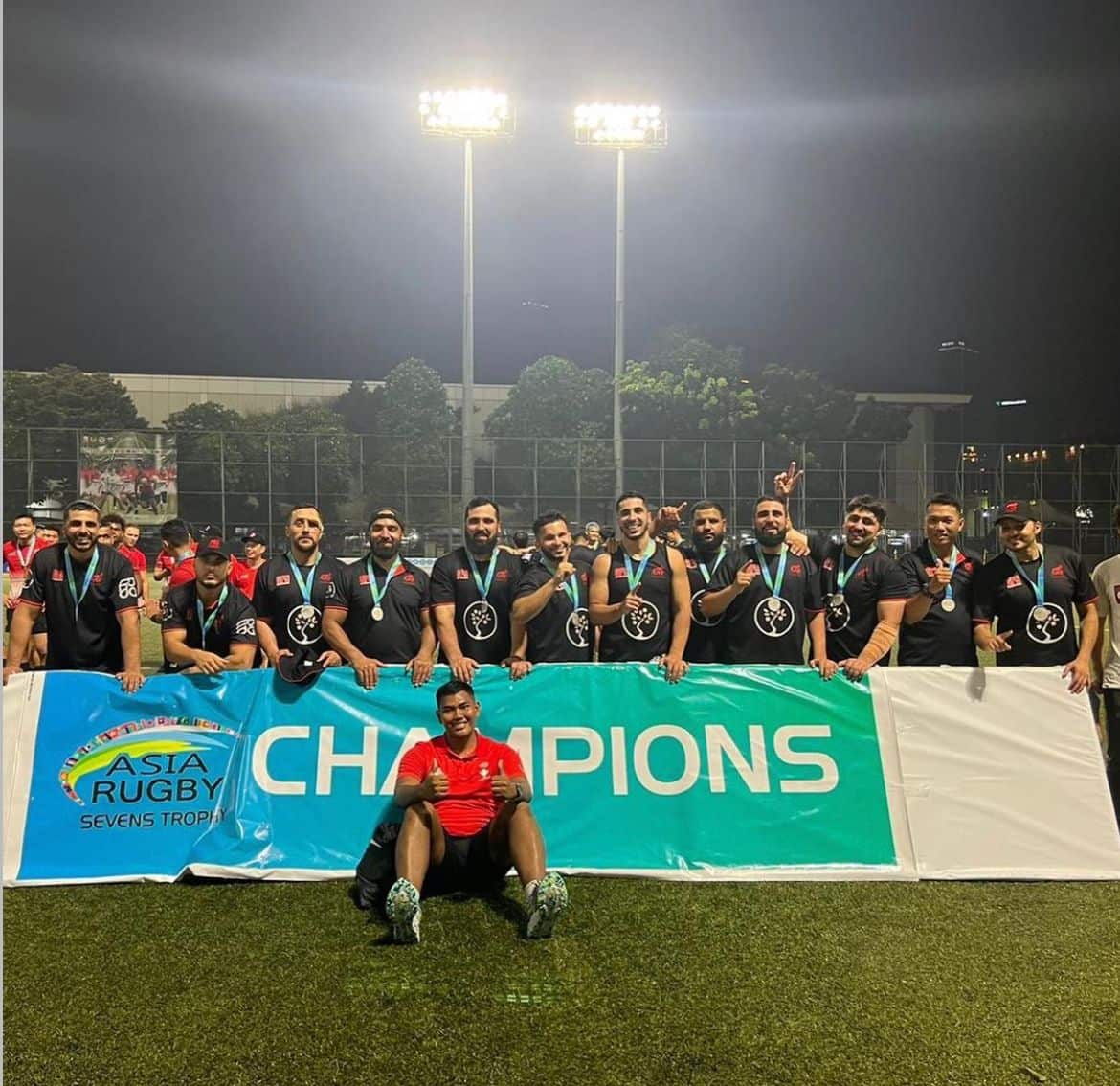 A line of men wearing silver medals smiling in front of a banner that reads 'CHAMPIONS' 