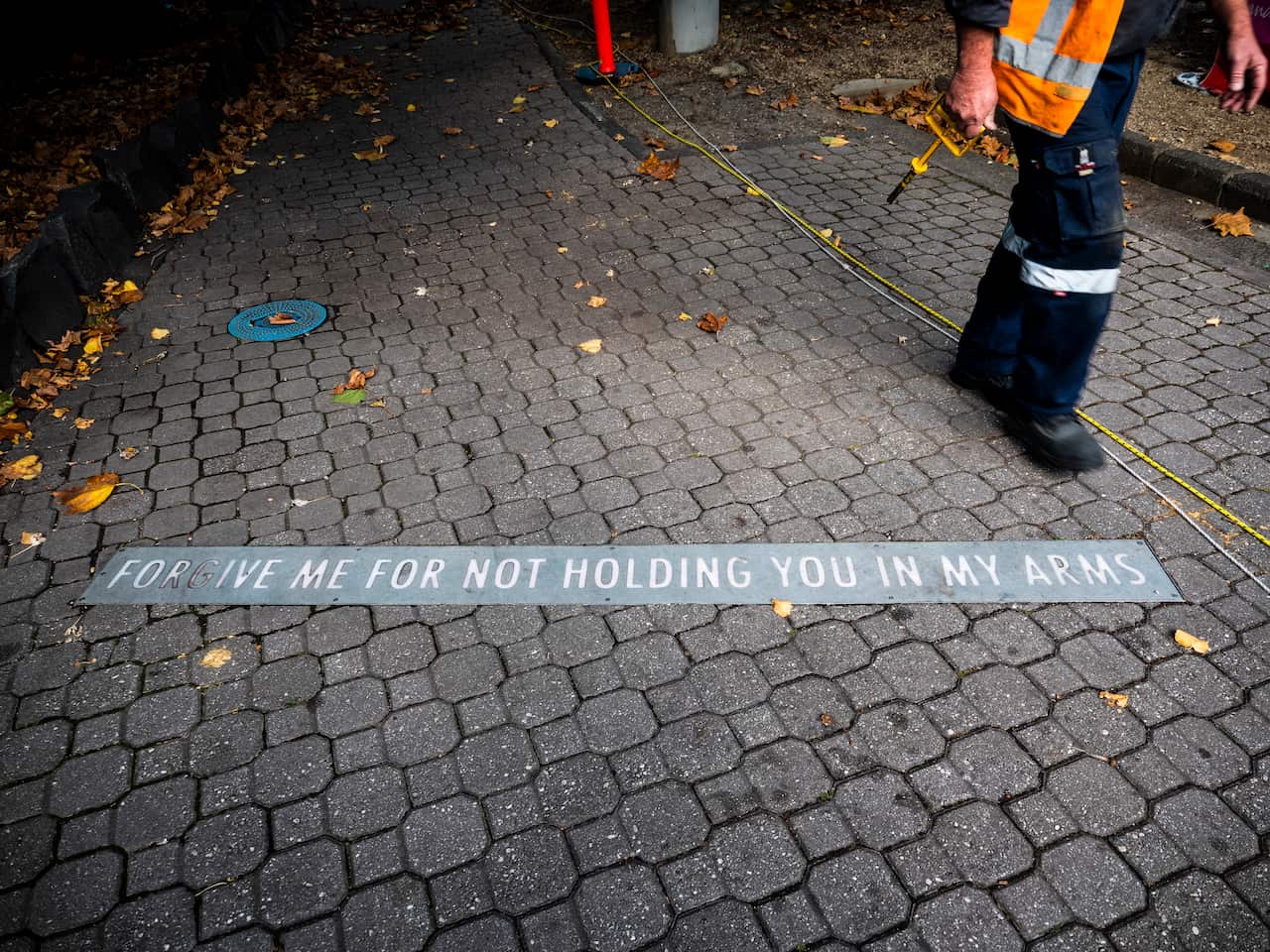 Worker walking past lettering on the ground saying 'forgive me for not holding you in my arms'. 