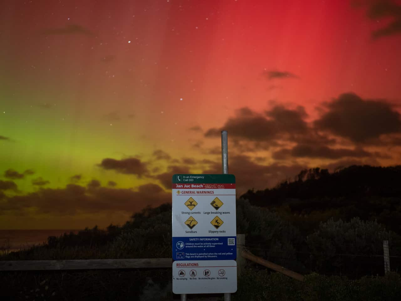 An aurora with colours consisting of green moving into red covers the night sky over a beach, with bush on the right side. A sign is in the middle warning beachgoers about rocky conditions