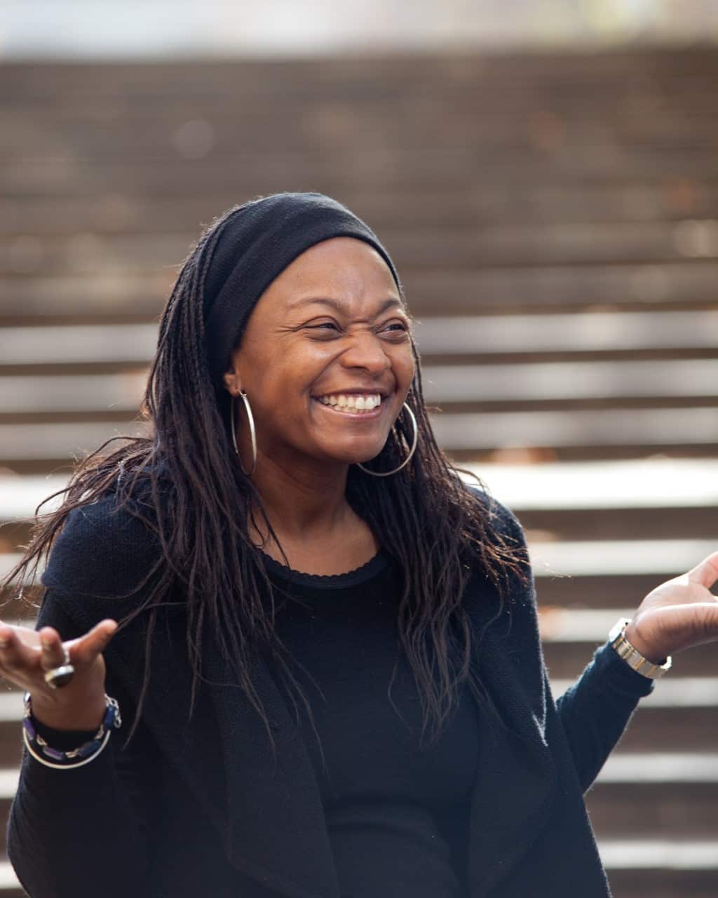 A woman dressed in black smiling while outside. She is holding both hands up with her palms out. 