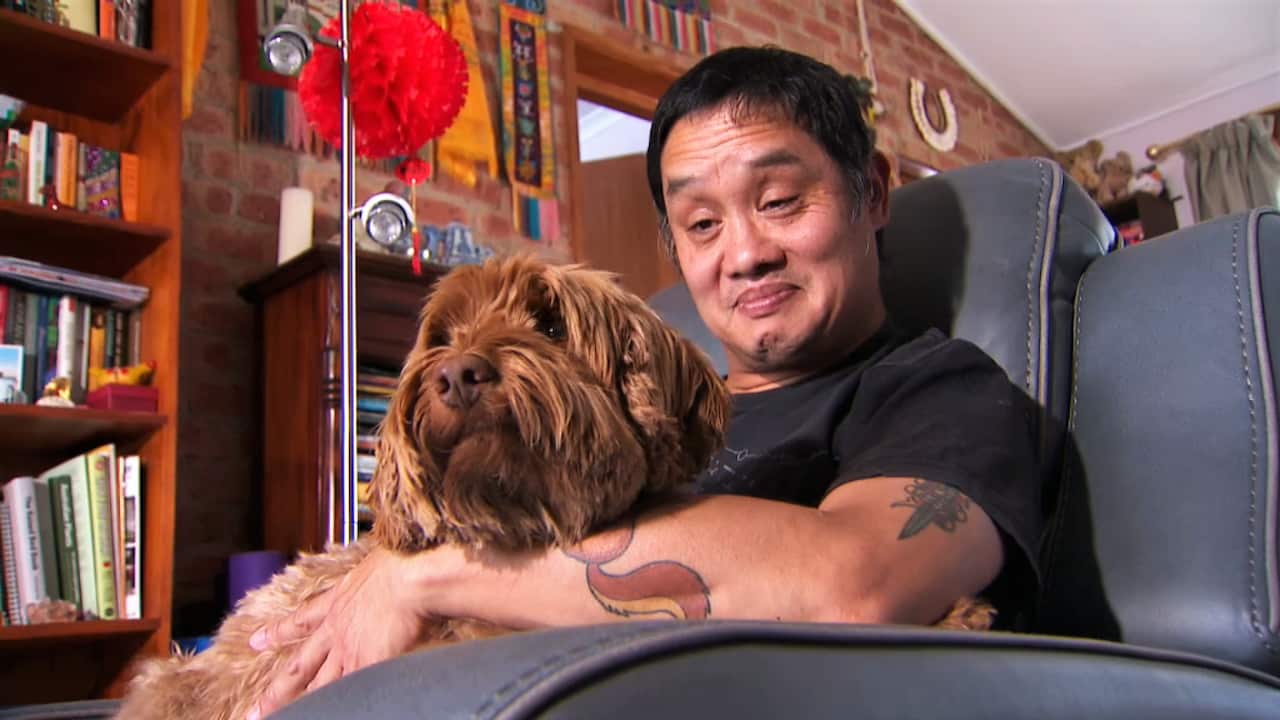 A man with tattoos on his left arm sitting in a black armchair holding his brown dog.