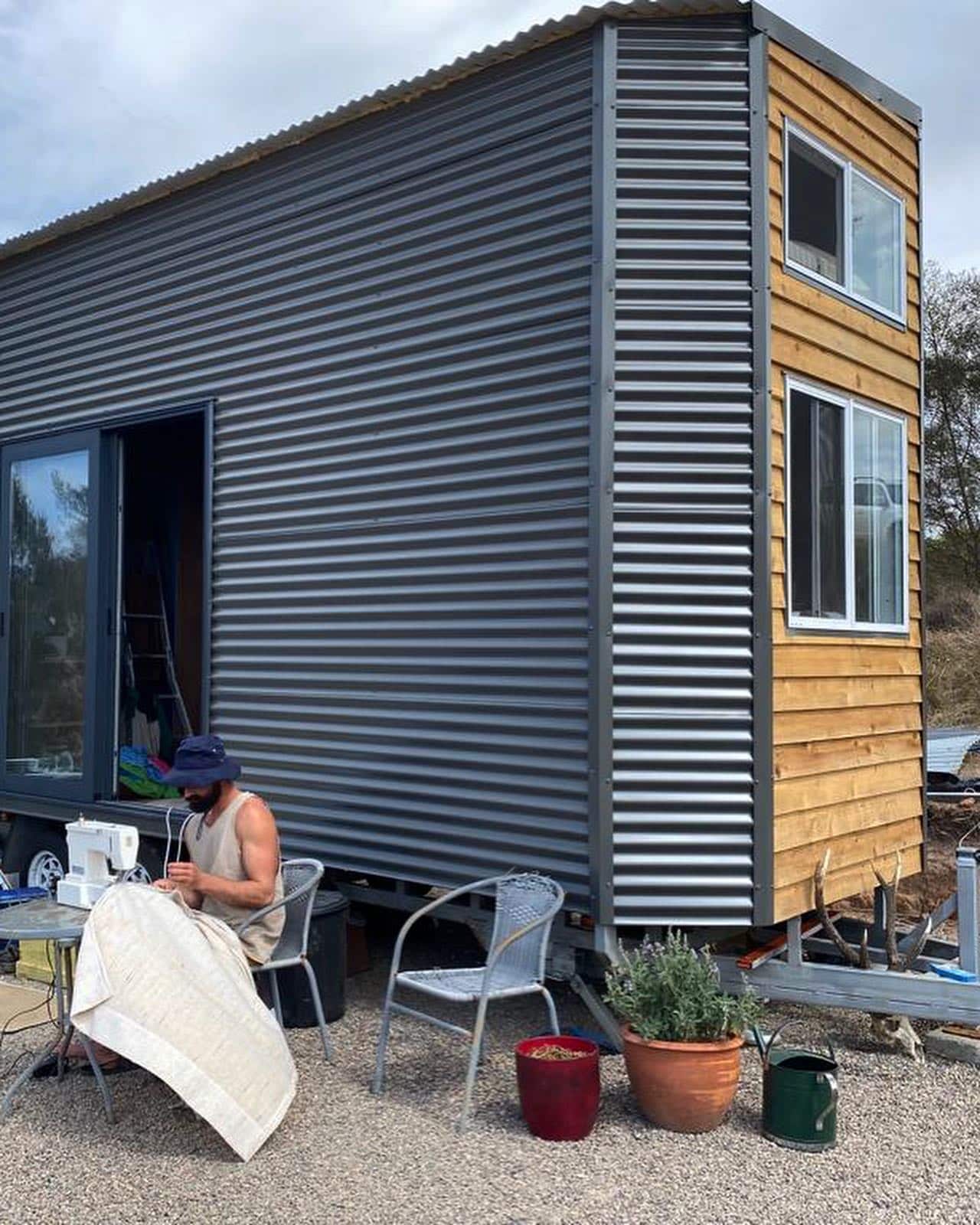 A man is sewing a curtain on a table outside a small home 
