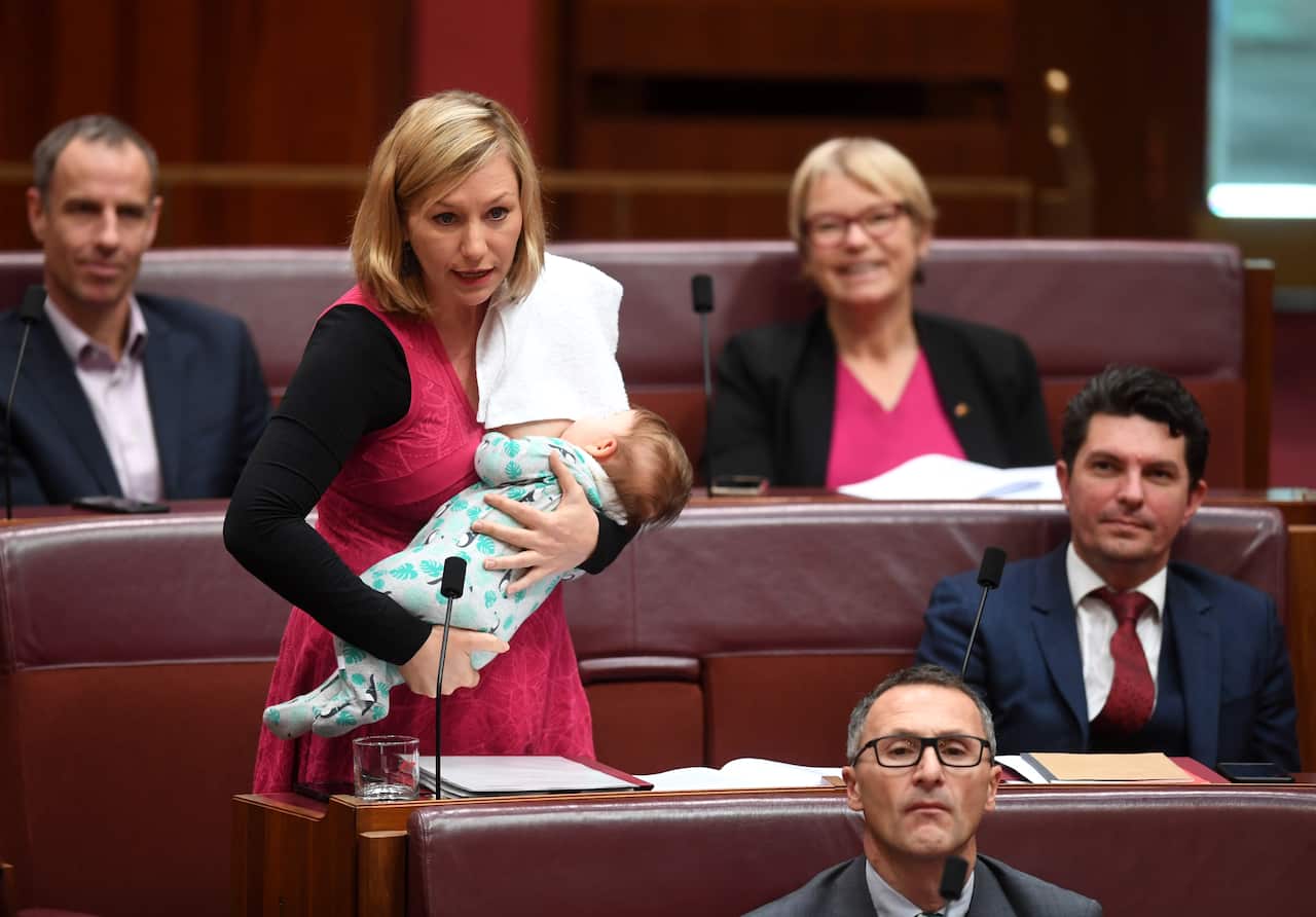 A woman in a red dress breastfeeds a baby in a senate chamber during a proceeding.