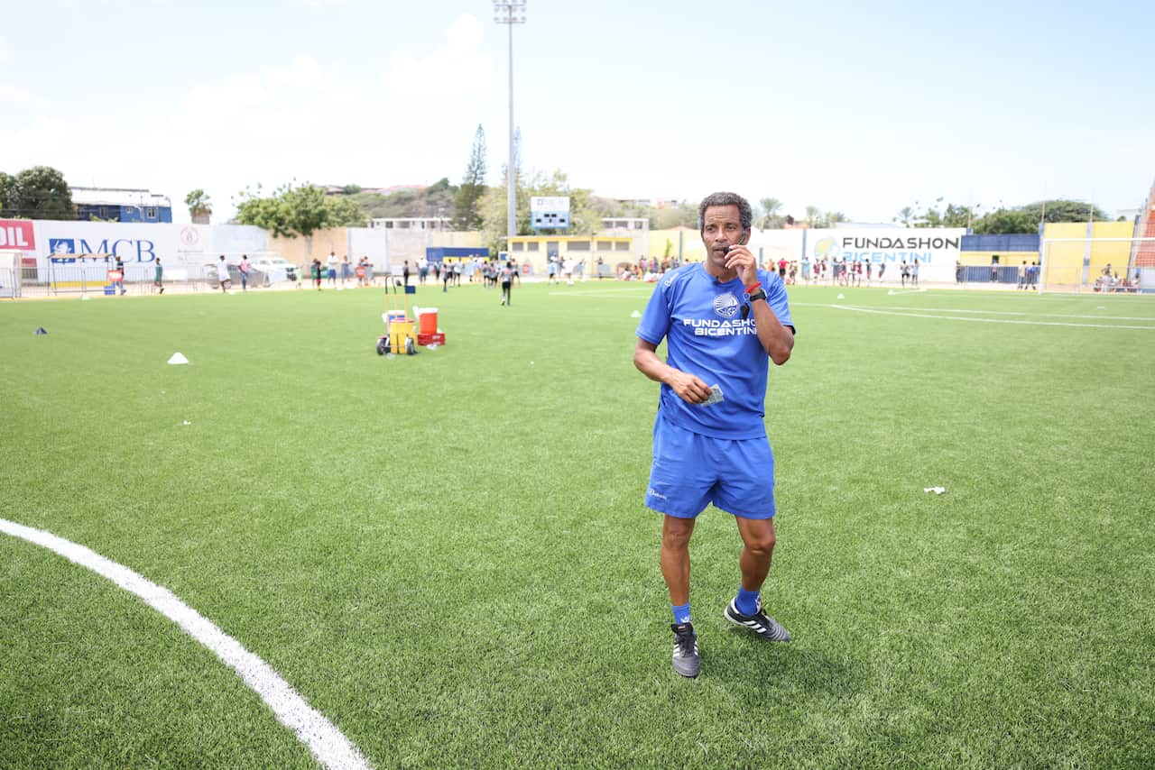 A man in a blue football uniform stands on a football pitch, speaking into the microphone of a headset. 