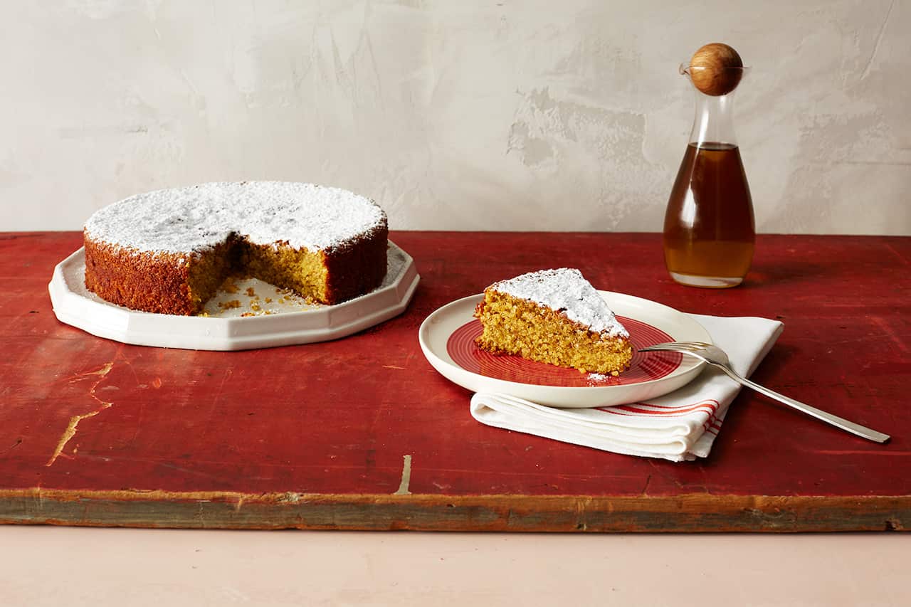 A round, low yellow cake dusted with icing sugar sits on a white plate on a red board. A slice of cake on a red and white plate also sits on the board, with a fork propped on the plate edge and a napkin underneath.  