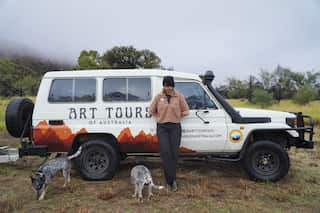 A woman leans against a white SUV parked in the middle of a green landscape. There are two dogs in the foreground.