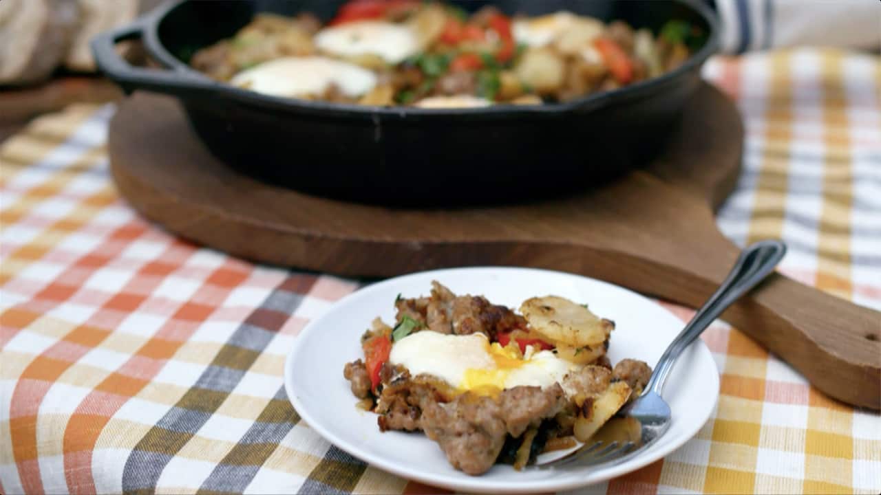 A black cast iron pan sits on a wooden paddle on a bright orange, yellow and grey checked cloth. It holds a hearty egg, sausage and potato bake. A portion has been scooped out and sits on a white plate in front of the pan, with a fork. 