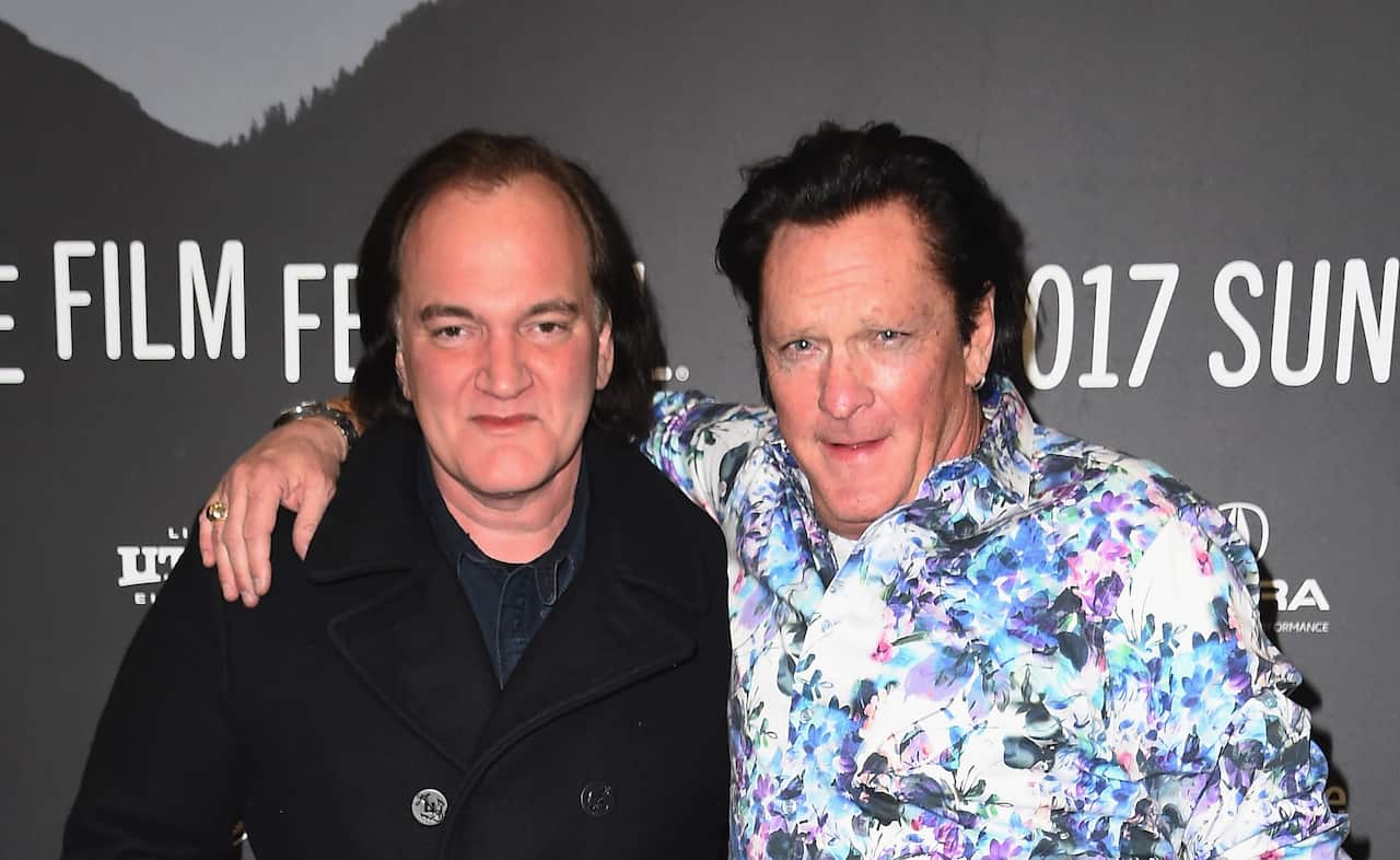 A man wearing a black blazer stands next to a man wearing a floral coat, with his arm draped around the other's shoulder.