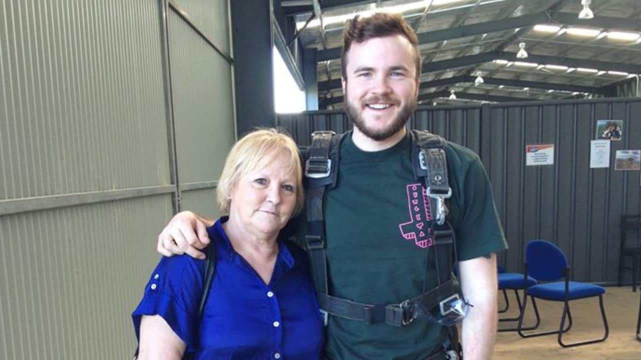 a middle aged woman in a blue top stands with a man in his early 20s who is geared up with a skydiving harness, inside a hanger