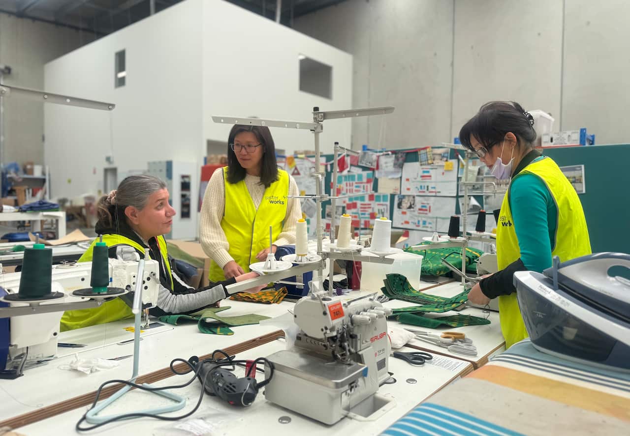Three women wearing hi vis vests are talking as they work inside a manufacturing hub with sewing machines in front of them.