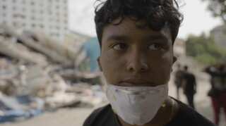 A close-up shot of a young man wearing an N95 mask on his chin with the rubble of a collapsed building behind him.