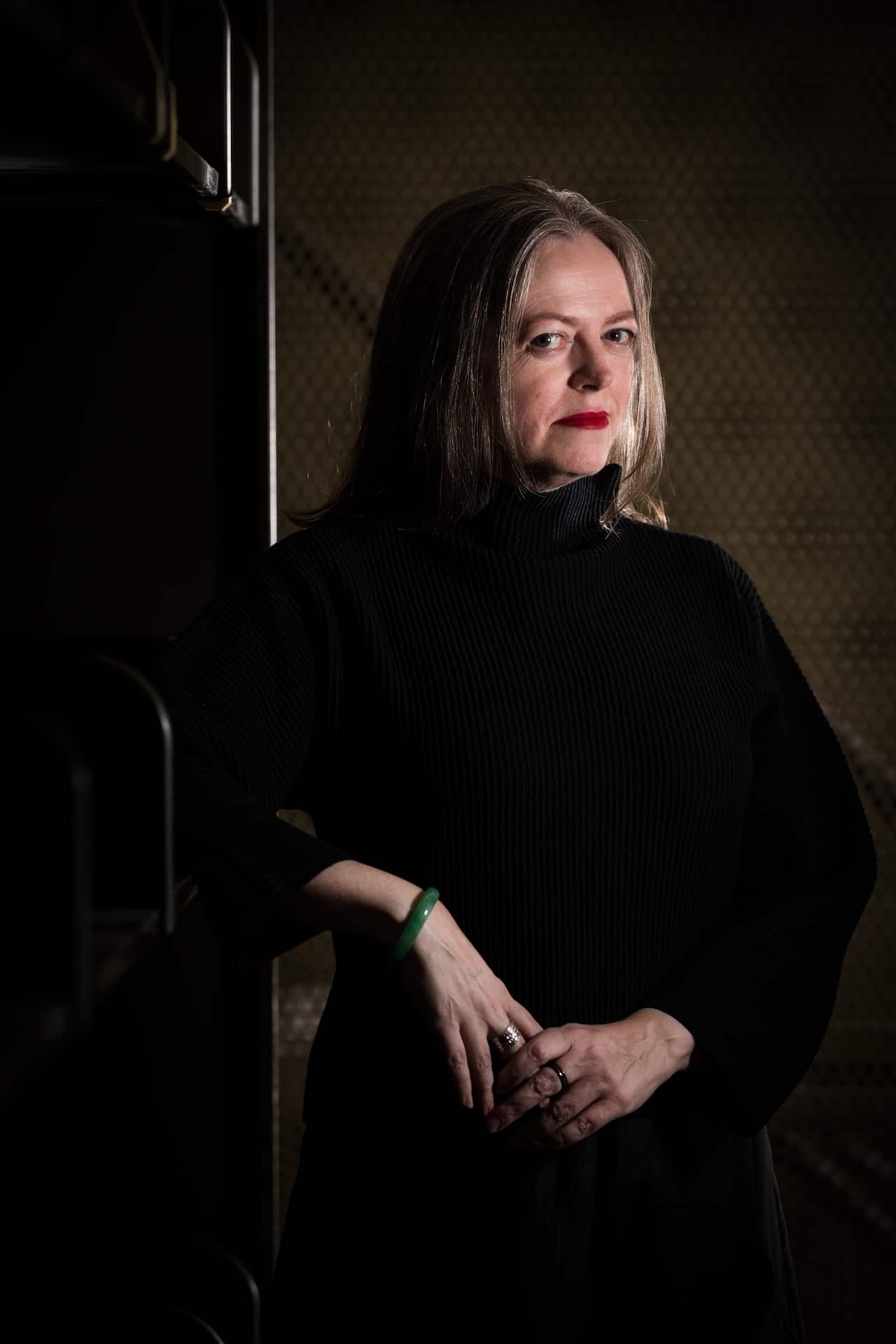 A woman wearing a black dress, standing against a dark background.
