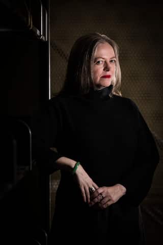 A woman wearing a black dress, standing against a dark background.