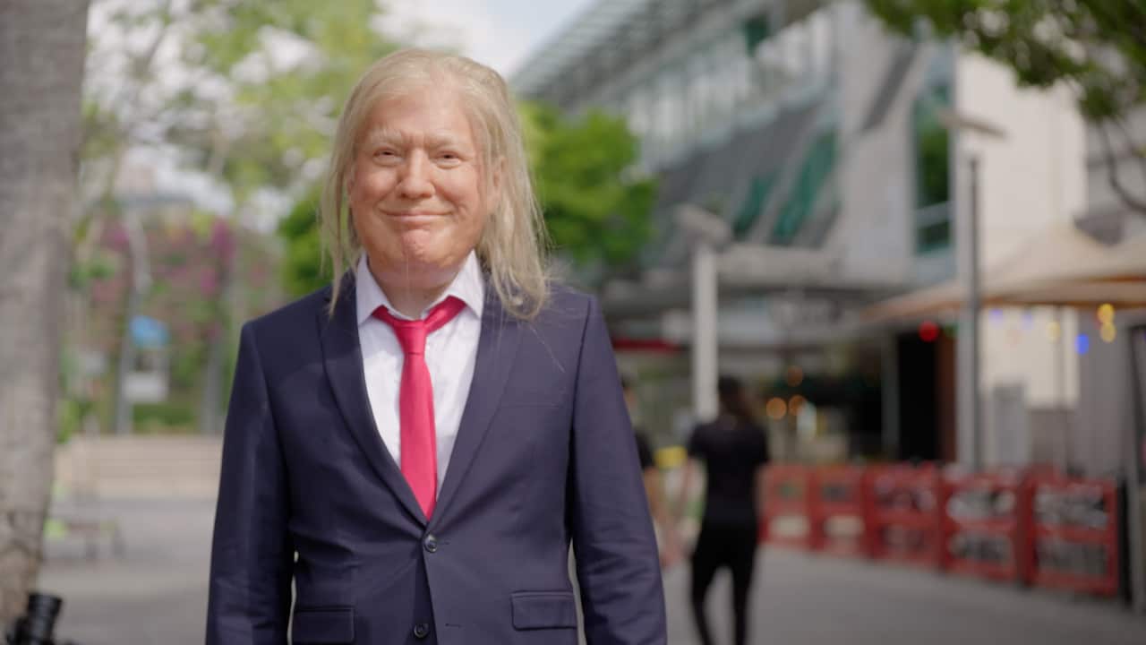 A man wearing a Donald Trump mask and a blue suit with a red tie is walking through the city of Brisbane