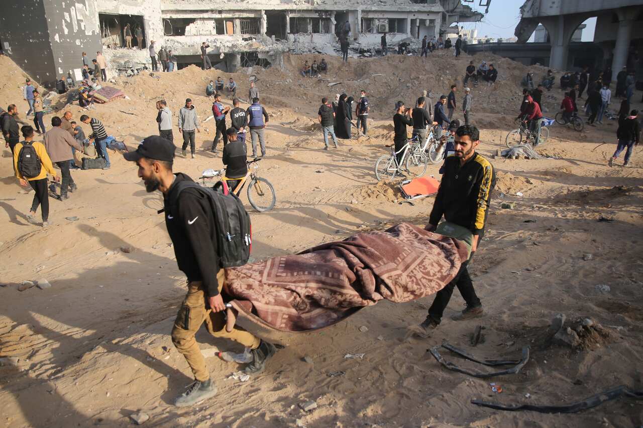 Two men carry a body wrapped in blankets on a stretcher amongst rubble
