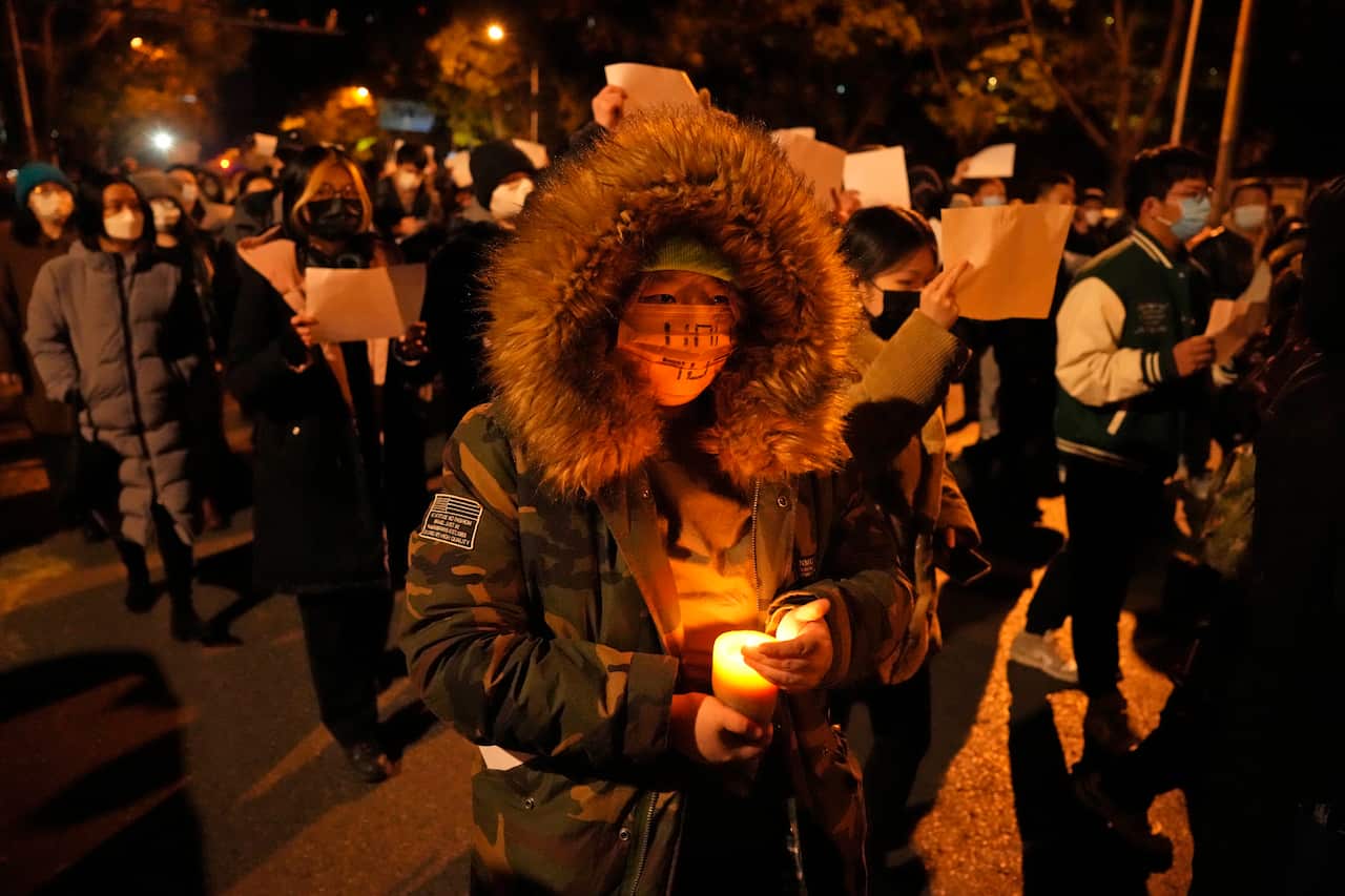 A man holding a candle and wearing a mask attending a protest against COVID-19 restrictions in China during the evening. Surrounding him are other protesters also wearing masks, holding up blank papers. 