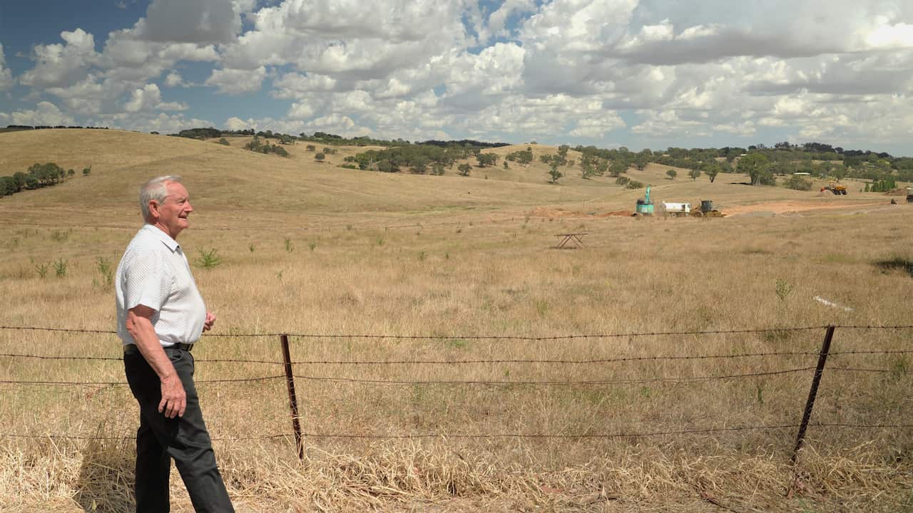 Older Caucasian man with white hair wearing a pale shirt and dark pants, walking along a fence line with vast rolling hills in the background, dotted with green trees and a blue sky speckled with fluffy white clouds. In the middle distance to the right is some heavy machinery undertaking construction work.