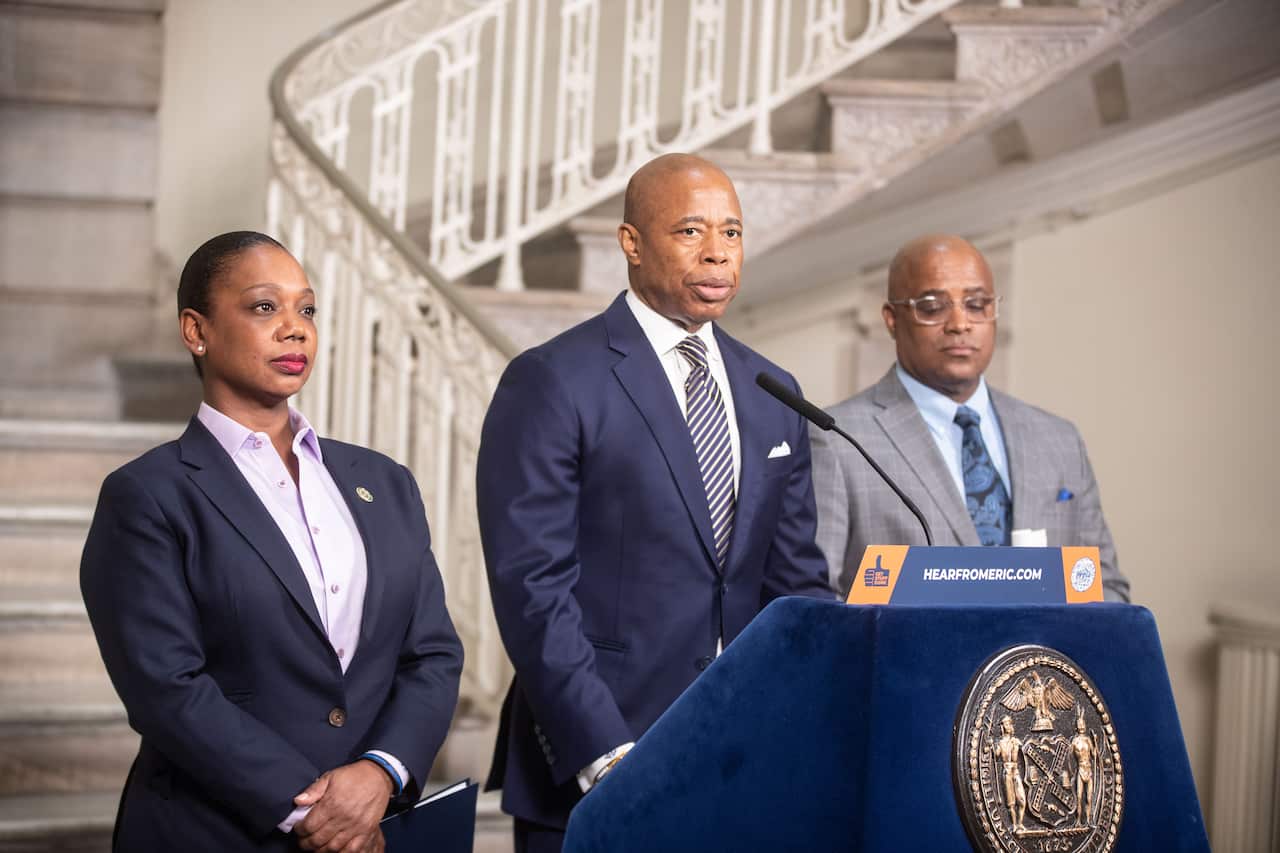 A man in a suit speaks from behind a blue lectern. There is a woman standing to his right and a man standing to his left.