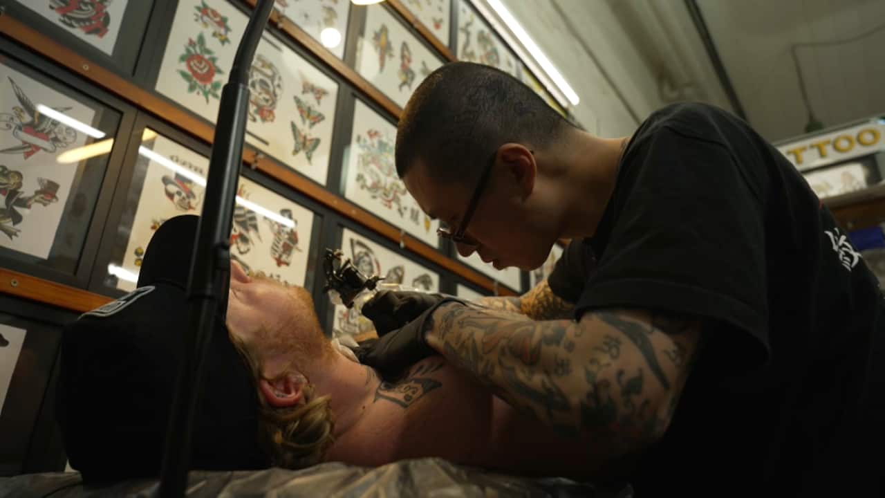 A man in a black T-shirt tattoos a man lying on a table in a tattoo parlour decorated with wall-to-wall framed pictures of tattoo designs.