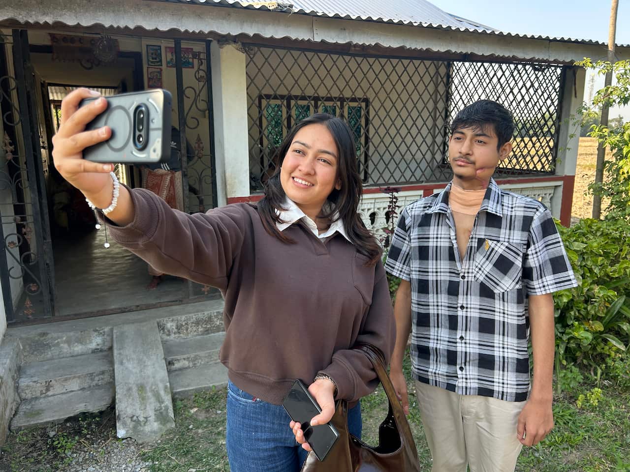 A smiling young woman takes a selfie with a young man, who has a bandage on his neck and a large scar on the left side of his face. 