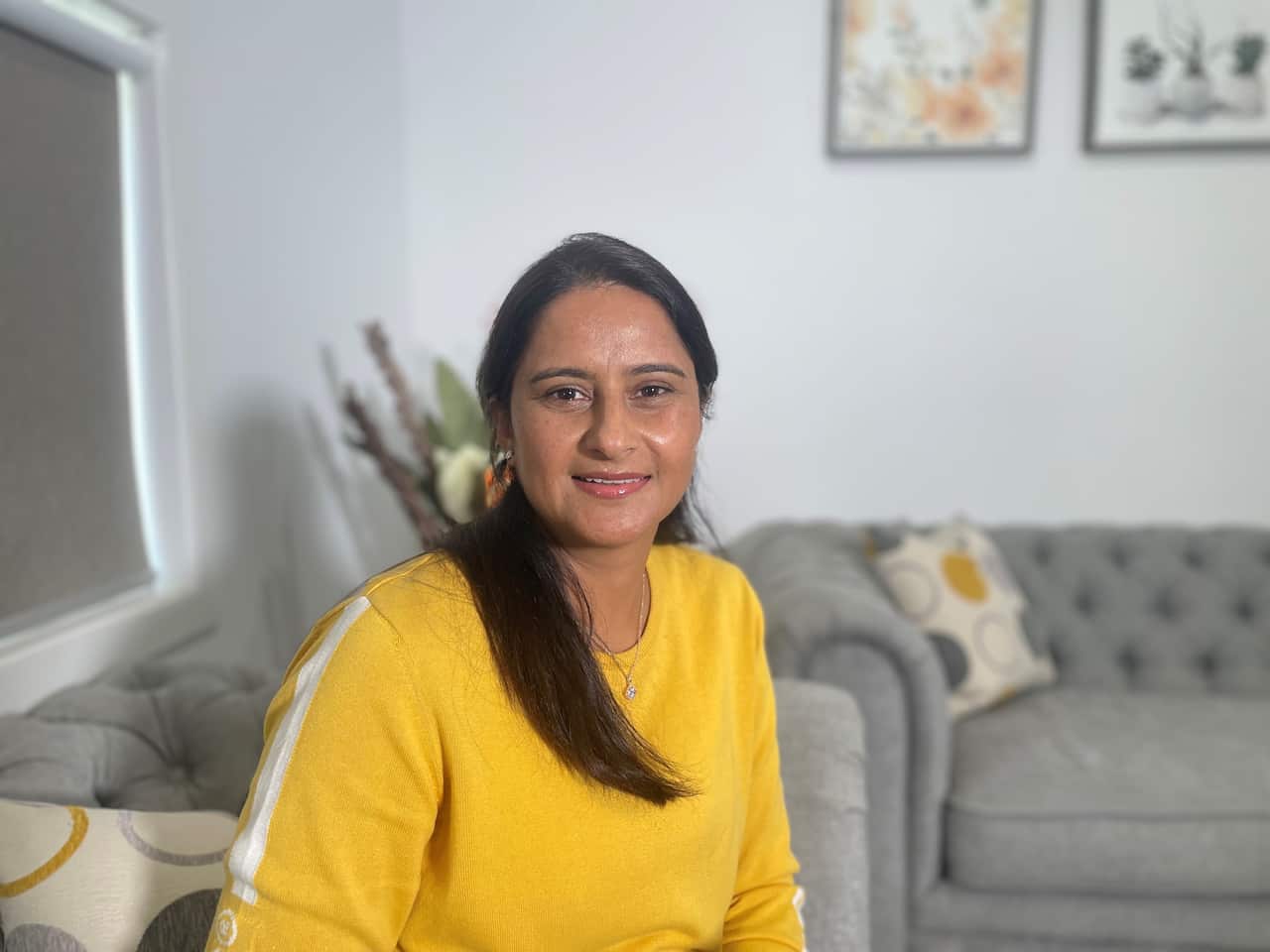 A woman in a long-sleeved yellow top in the room of a house smiles for the camera.