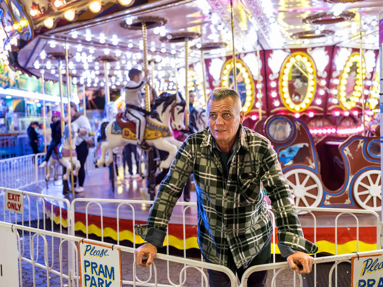 A man in a check shirt stands in front of a merry-go-round. 