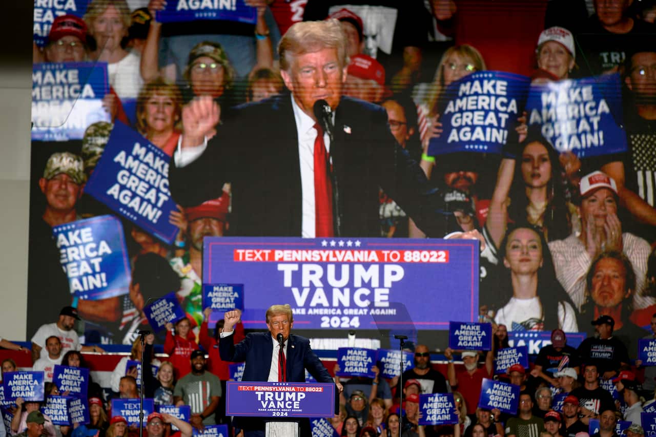 A man stands in front of a podium with 'Trump Vance' signs in the background