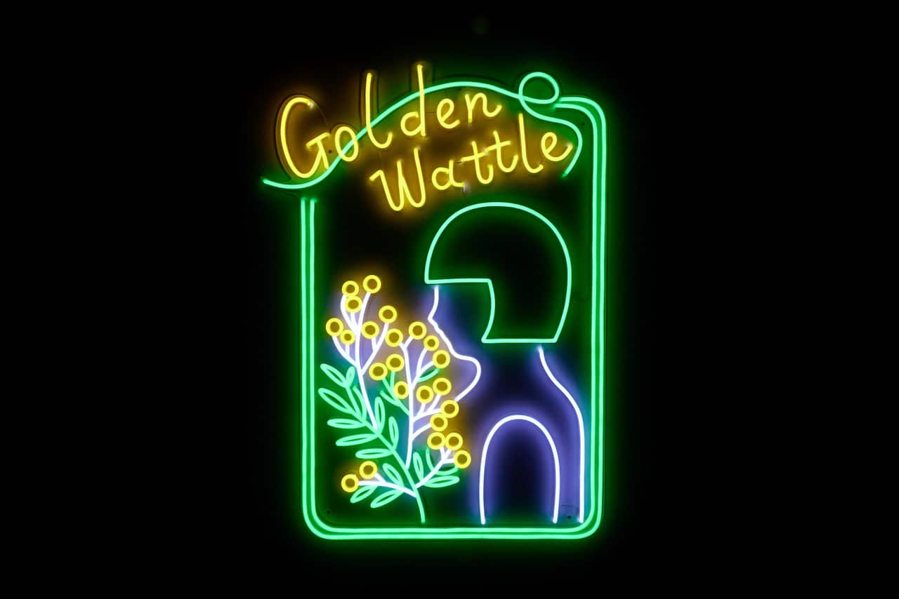 A neon green and yellow sign that reads "Golden Wattle" and depicts the figure of a young woman and a wattle branch.