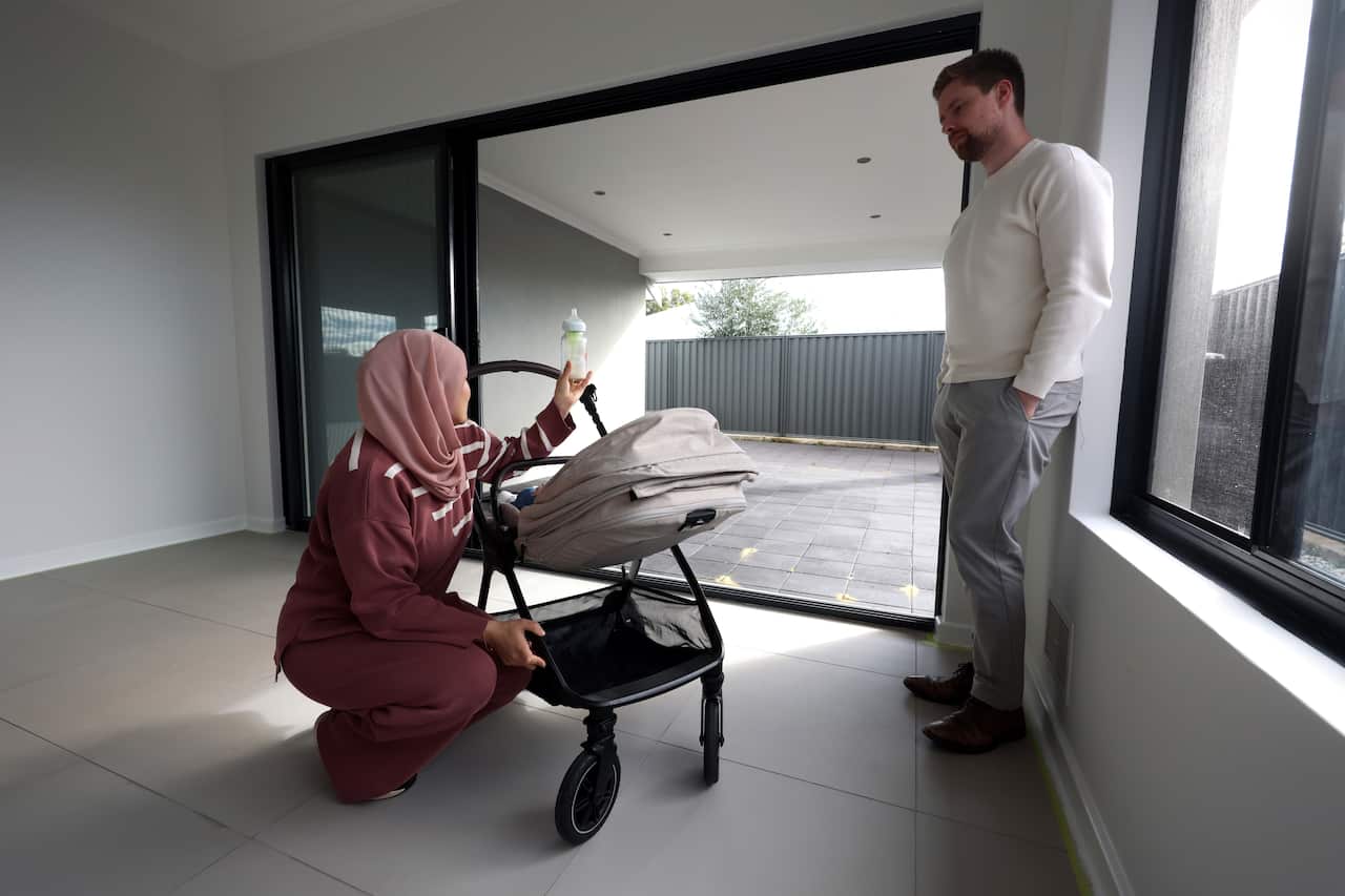Fatima Payman feeds her daughter a bottle at her home. Her husband stands next to them in an empty family room.